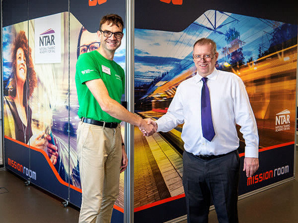 byran denby and simon rennie shake hands in front of a mission room arena display system