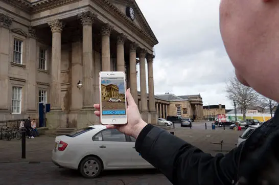 Image of a woman holding a phone showing Huddersfield train station, with the actual station in the background 